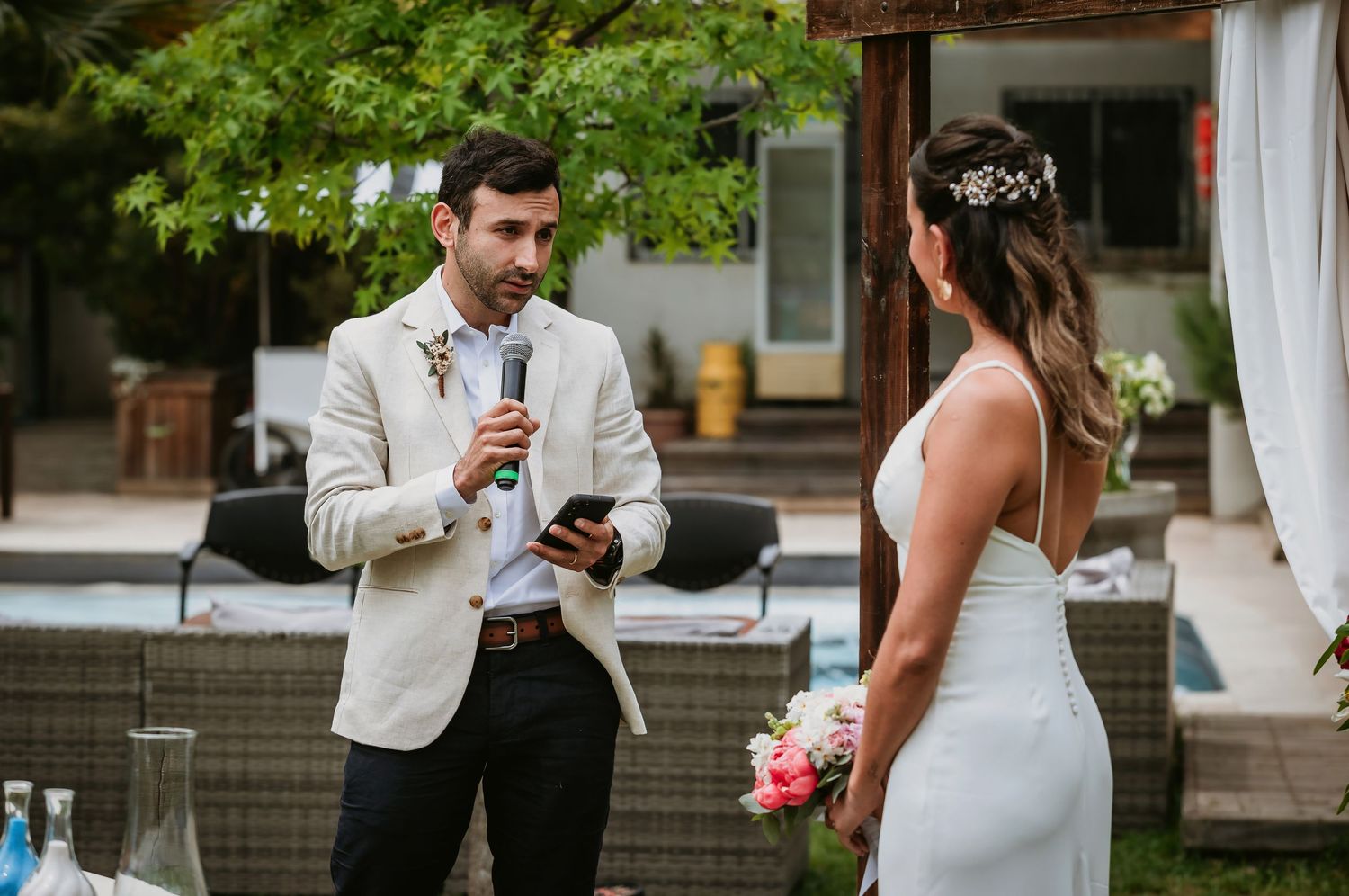 Groom in white blazer reads vows to bride holding bouquet during outdoor wedding ceremony.