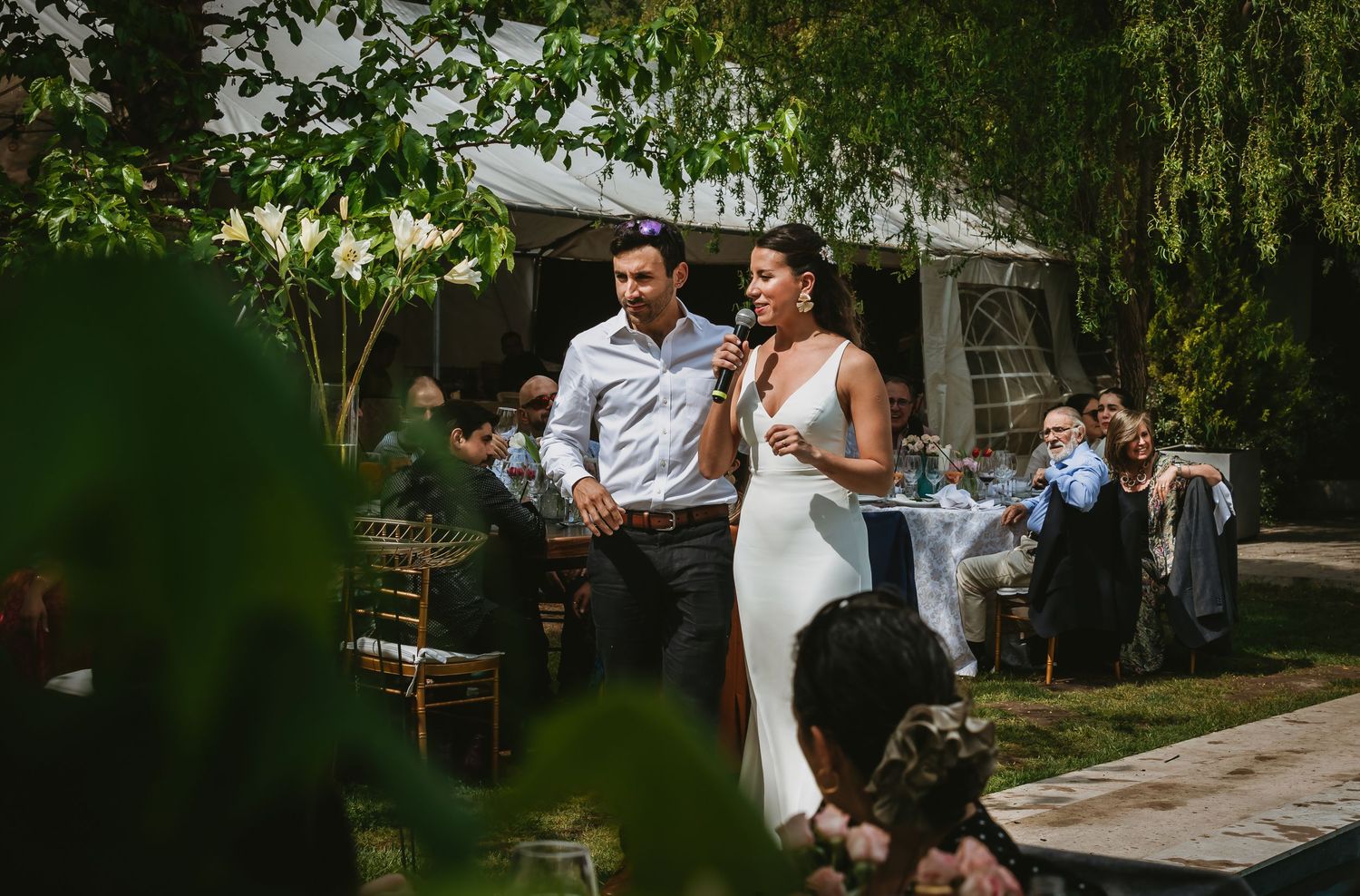Bride in white gown speaks into microphone beside groom at outdoor wedding reception surrounded by lush greenery.