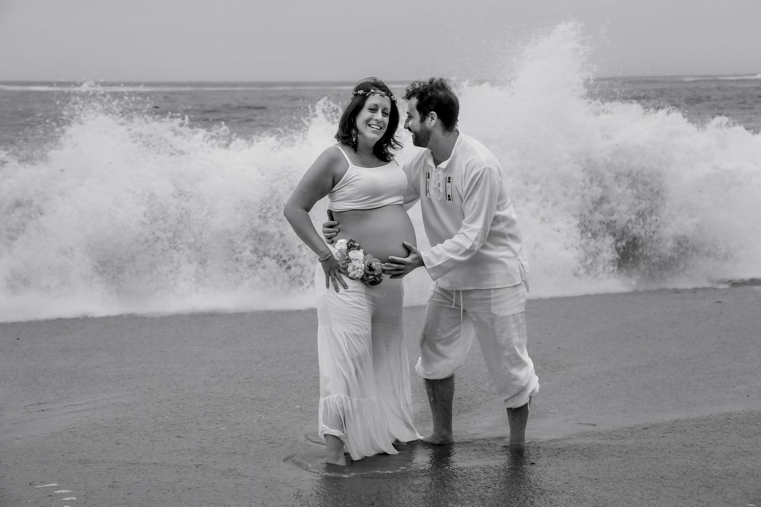 Black and white photo of a pregnant couple embracing on a beach with dramatic crashing waves behind them.