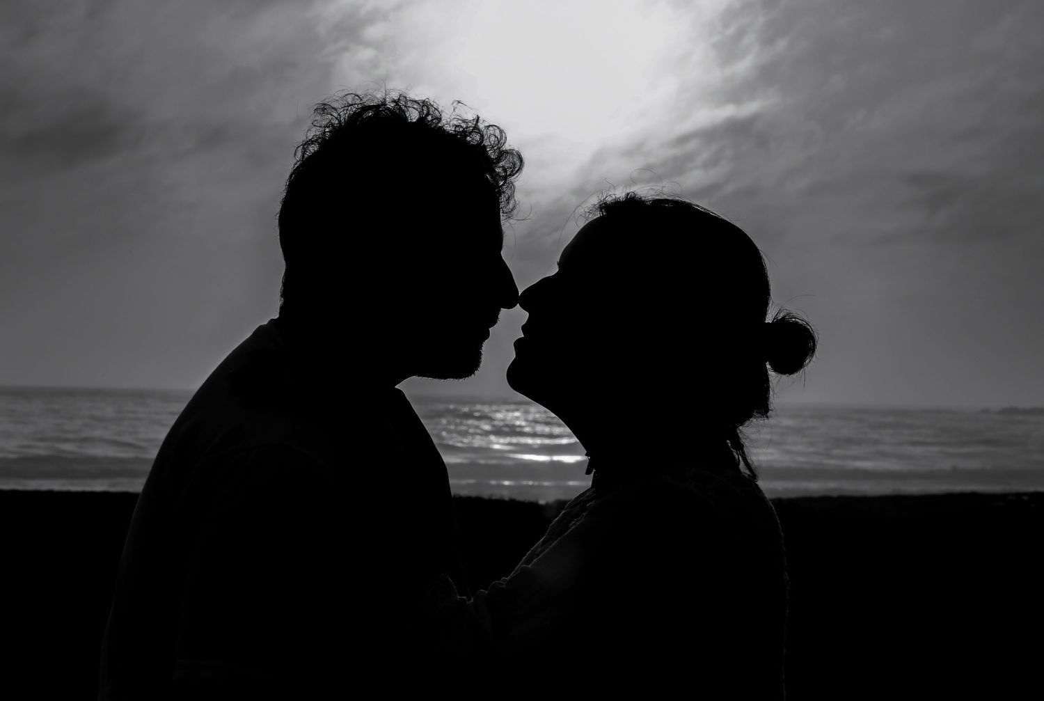 Black and white silhouette of a romantic couple about to kiss at sunset near the ocean.
