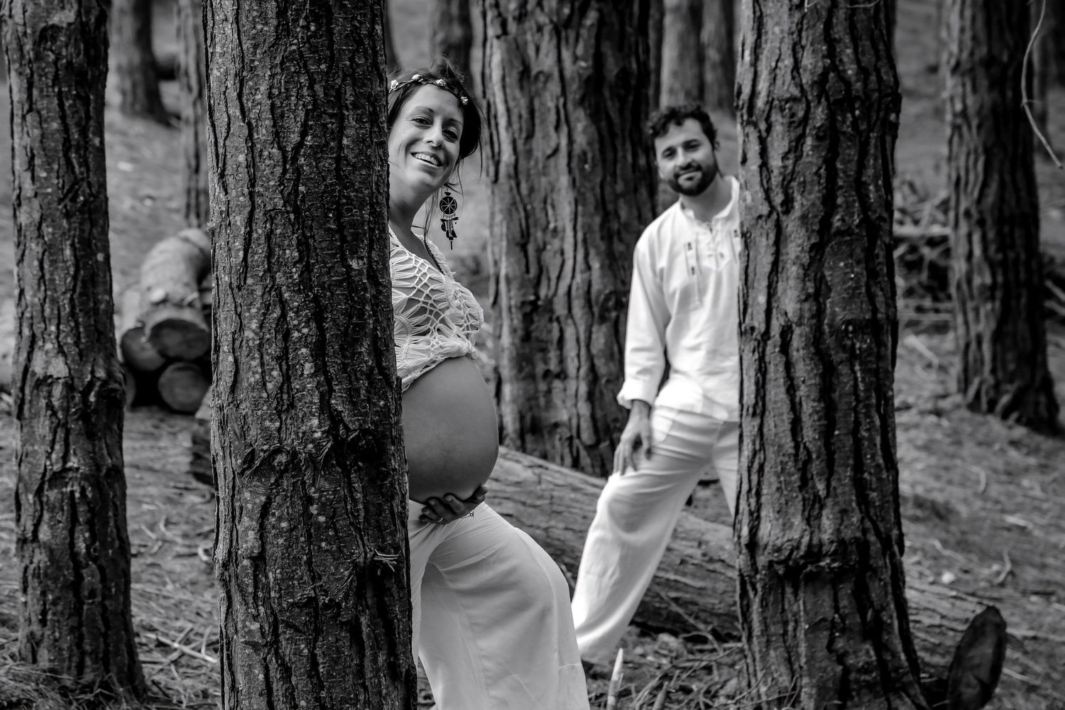 Black and white maternity photo of pregnant couple posing between forest trees, woman showing baby bump, man standing behind.