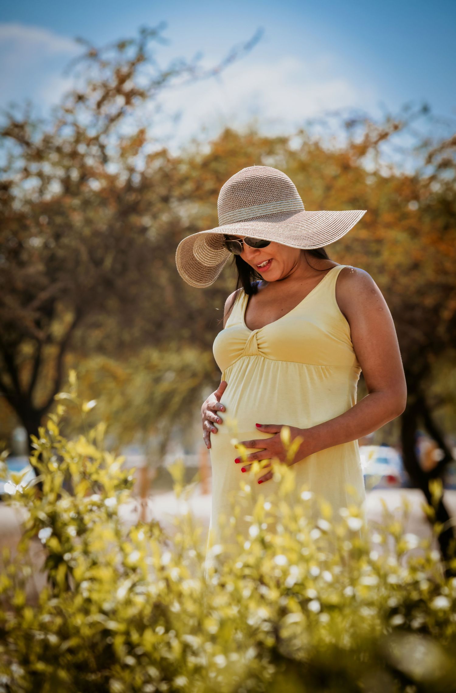 Pregnant woman in yellow dress and wide-brim hat cradling baby bump outdoors among green plants on a sunny day.