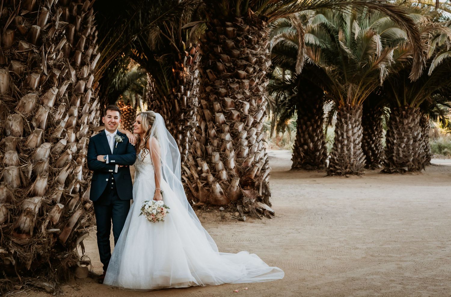 Wedding couple posing elegantly beneath a canopy of tall palm trees on sandy ground.