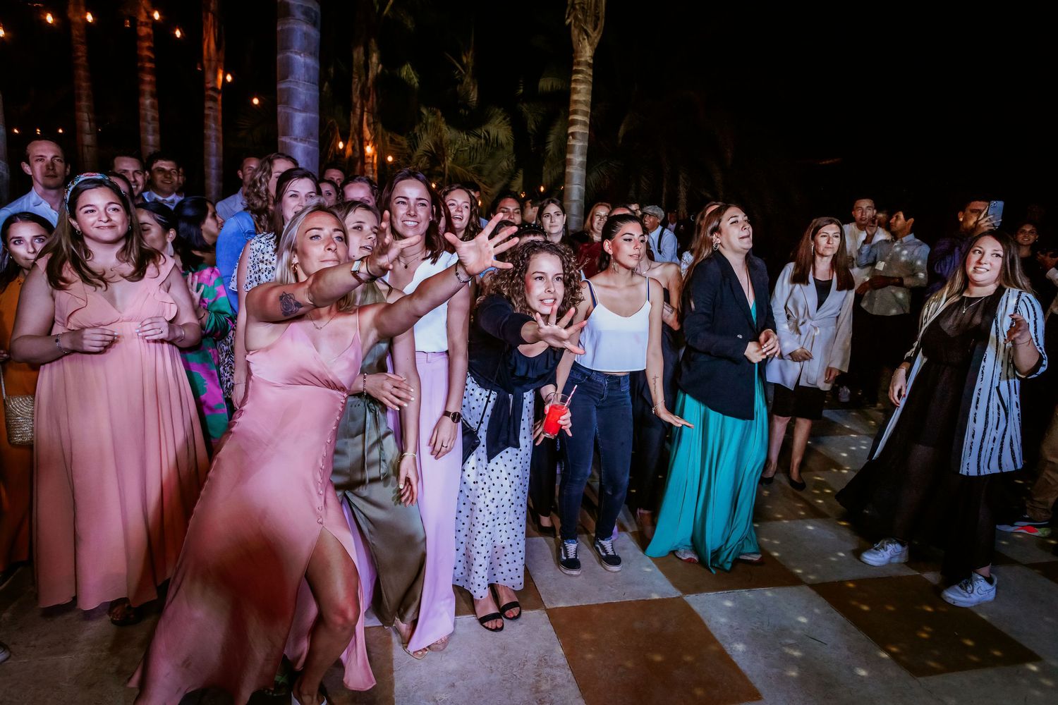 Excited wedding guests reaching for bouquet toss at outdoor evening reception under palm trees.