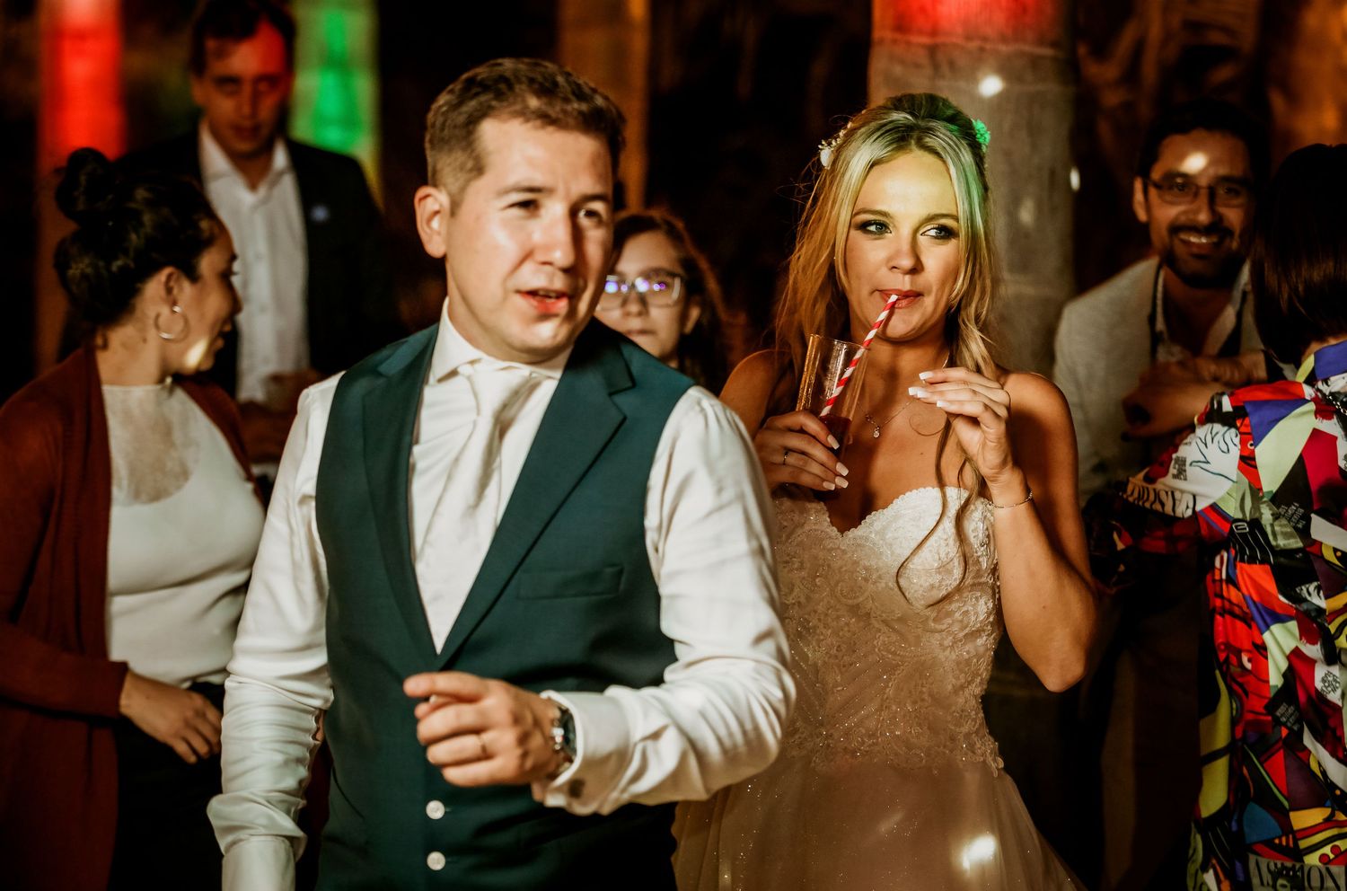 Bride holding champagne dances with guest in navy vest at elegant wedding reception.