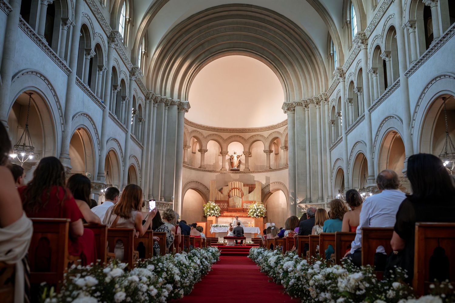 Wide view of packed church with red carpet aisle lined with white flowers during wedding ceremony.