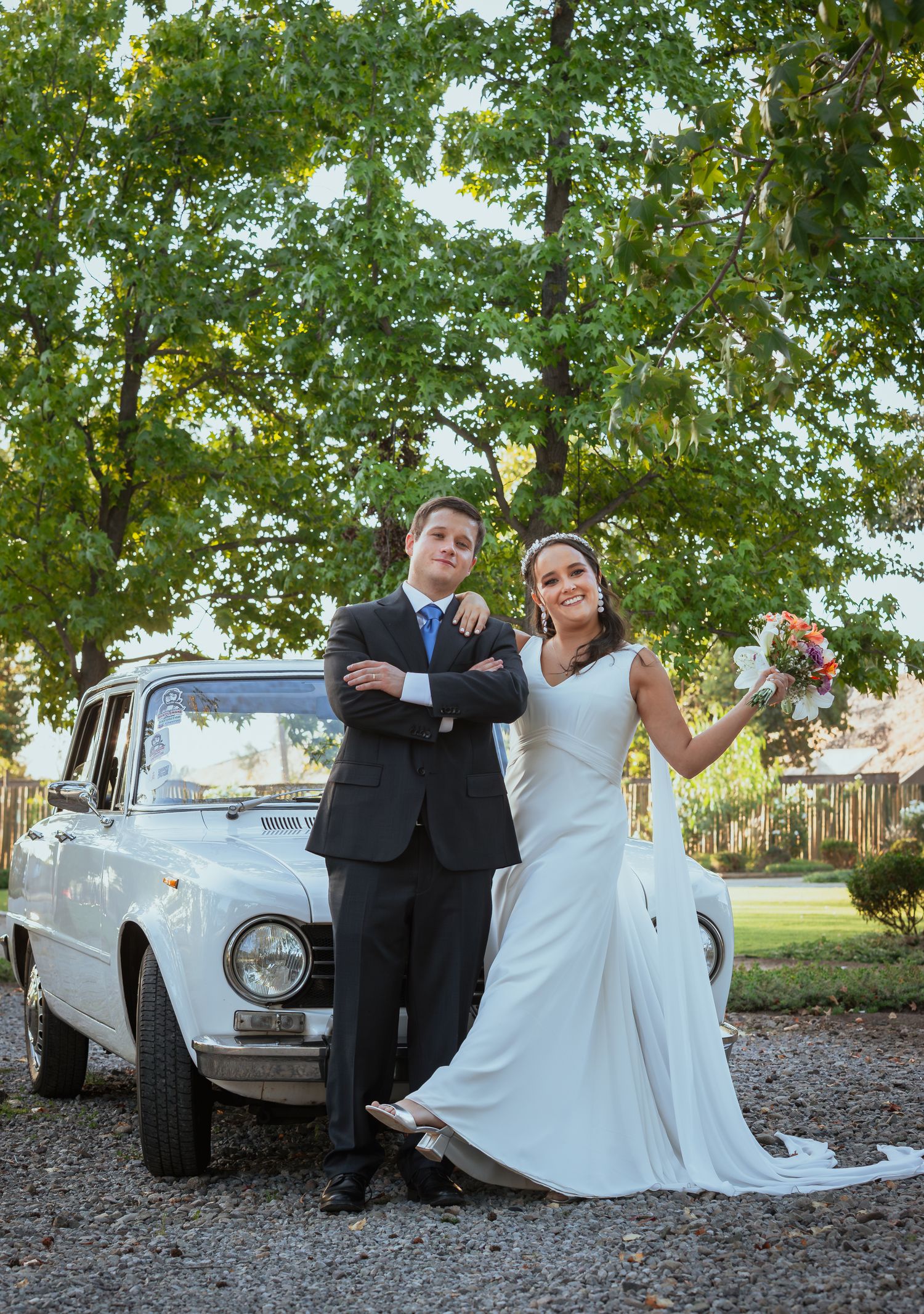 Bride and groom pose elegantly in front of classic white vintage car outdoors.