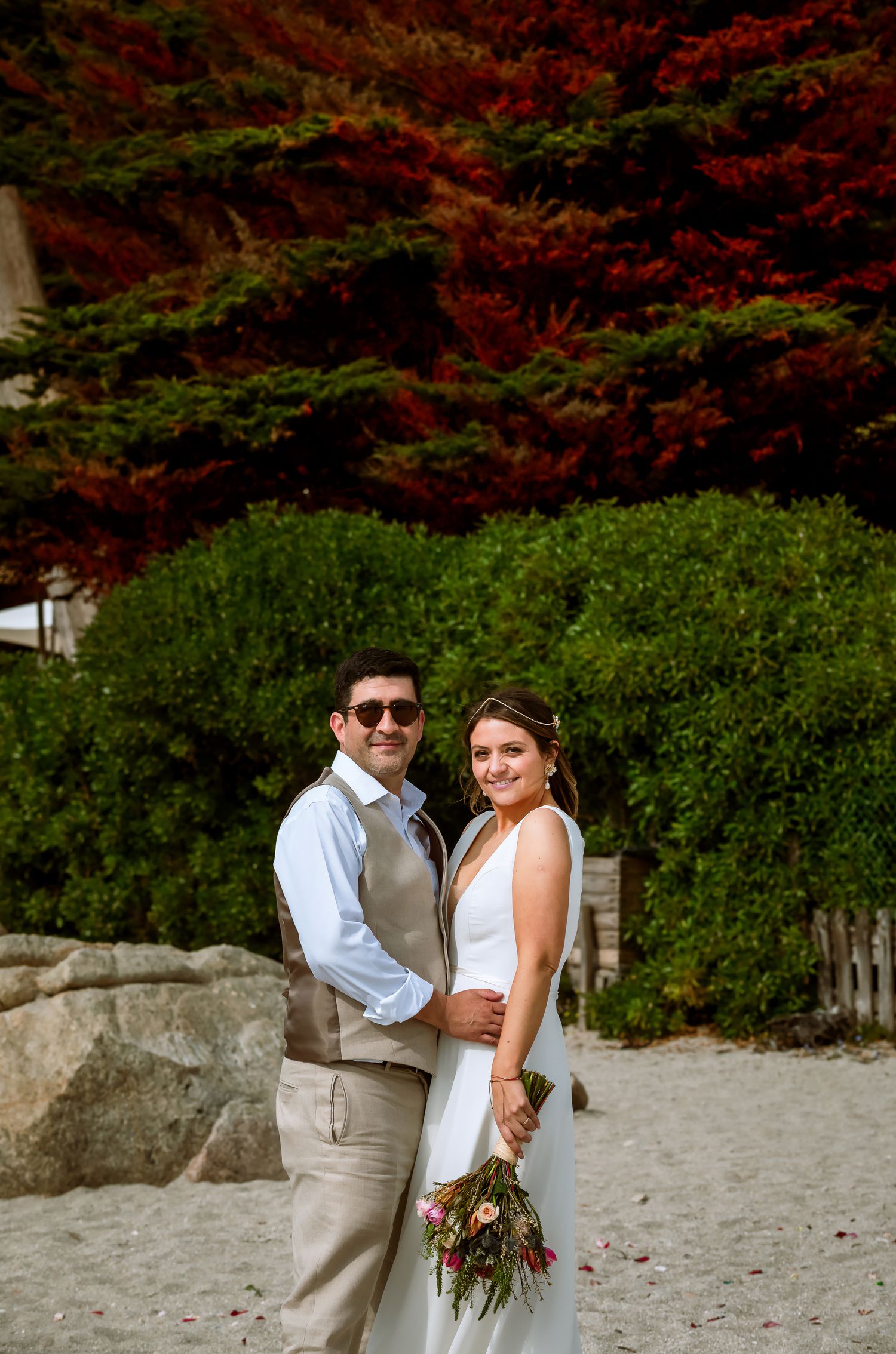 Wedding couple embracing on sandy beach, bride holding floral bouquet with vibrant red trees in background.