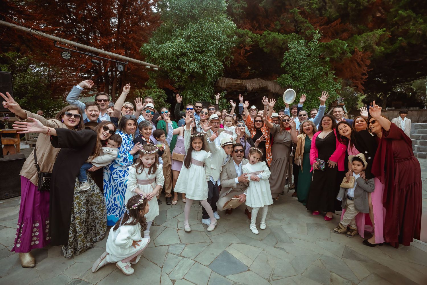 Large joyful wedding group photo with guests cheering outdoors among autumn trees.