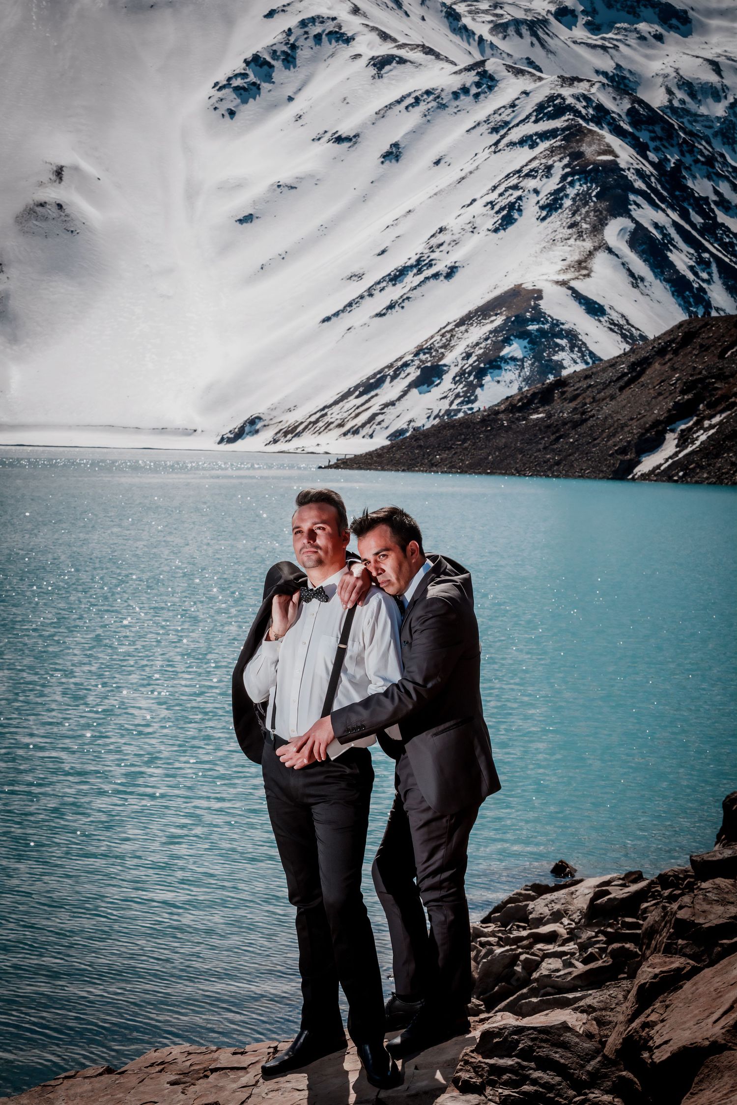 Two men in wedding suits embrace on rocky shore with turquoise mountain lake in background.