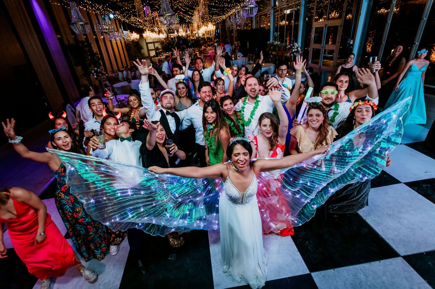 Bride with glowing LED wings poses with large group of wedding guests on checkered dance floor.