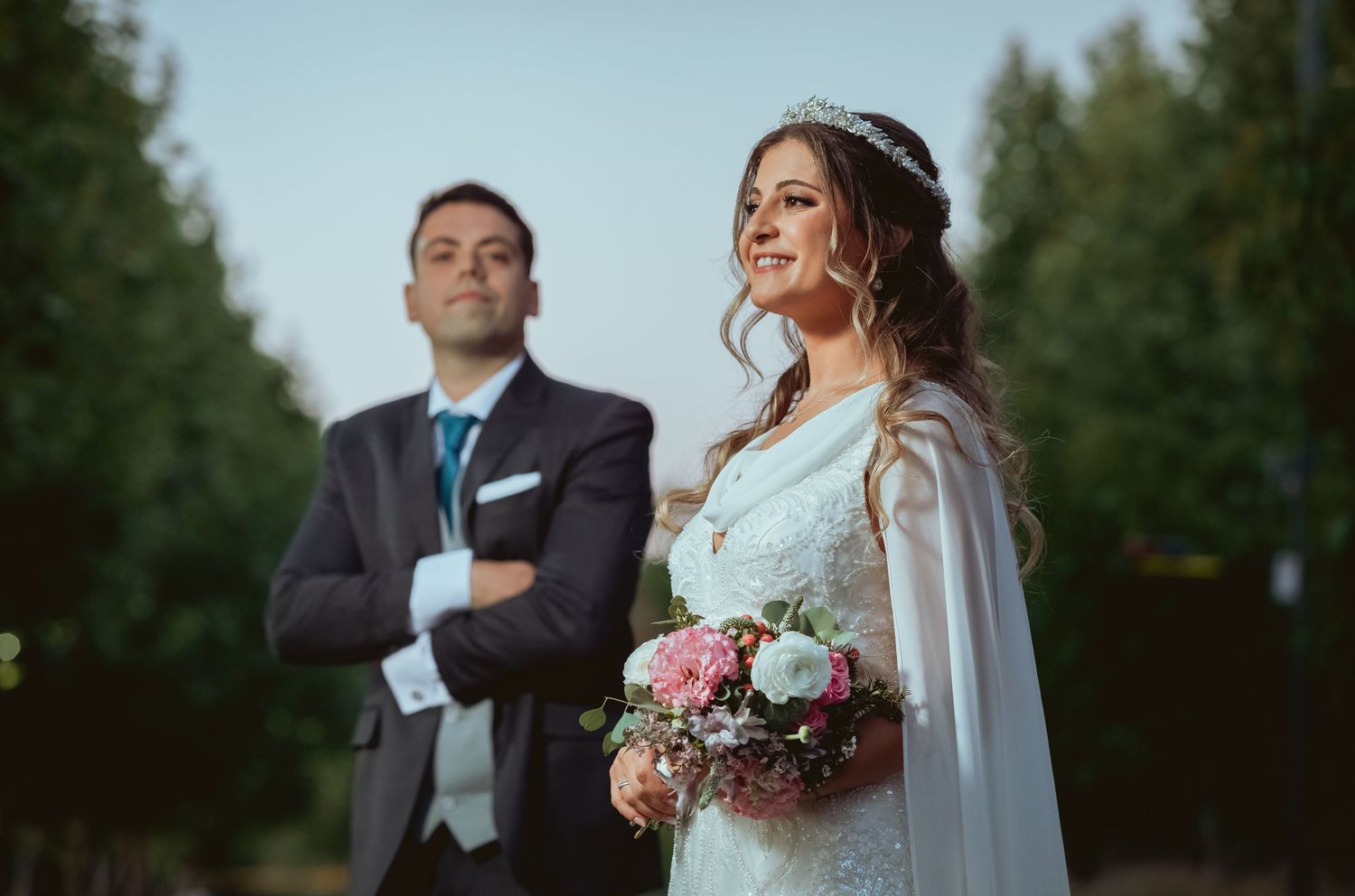 Bride in white gown holds bouquet while groom stands with crossed arms in a sunny outdoor setting.