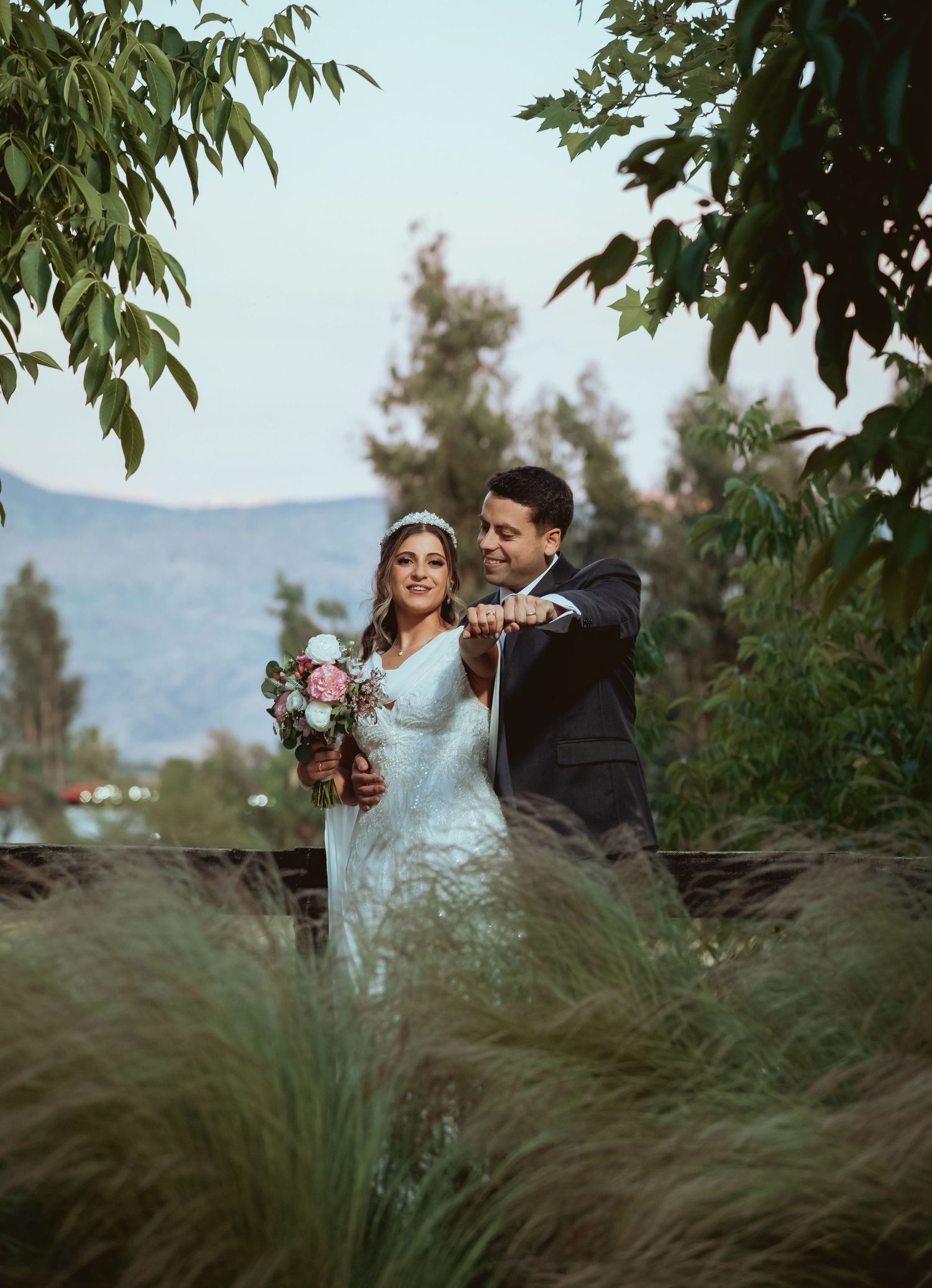 Bride holding pink bouquet stands with groom near a lake with lush greenery in the background.