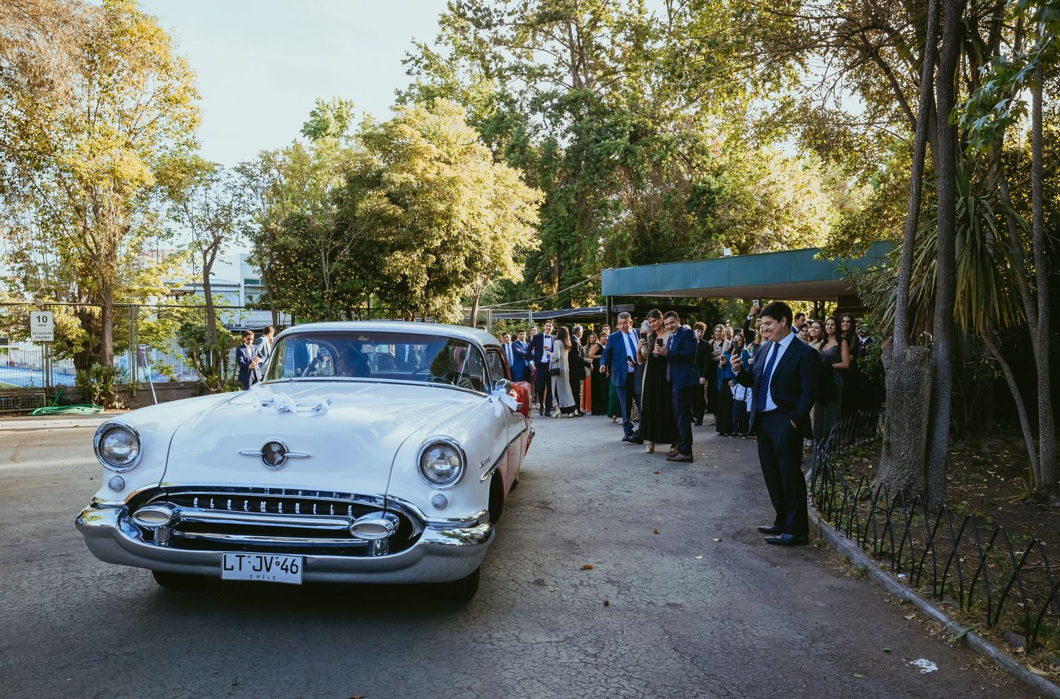 White vintage classic car at a wedding with elegantly dressed guests gathered outside amid lush green trees.