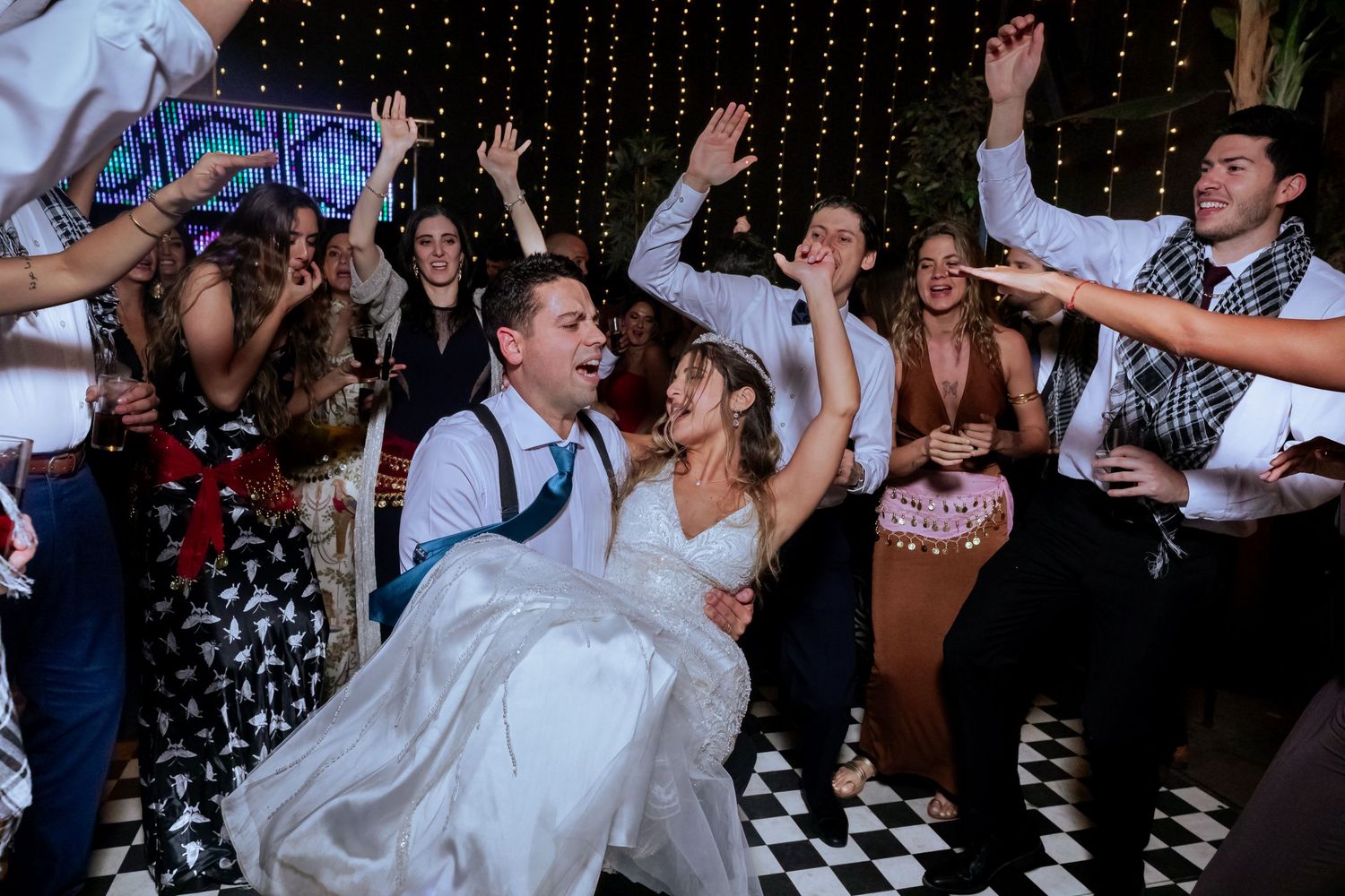 Bride and groom laugh and dance energetically with guests on a black and white checkered dance floor.