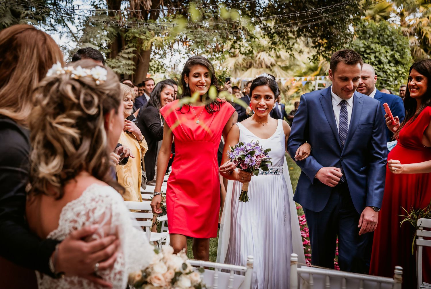 Wedding guests smile as bride walks down aisle in outdoor garden ceremony surrounded by lush greenery.
