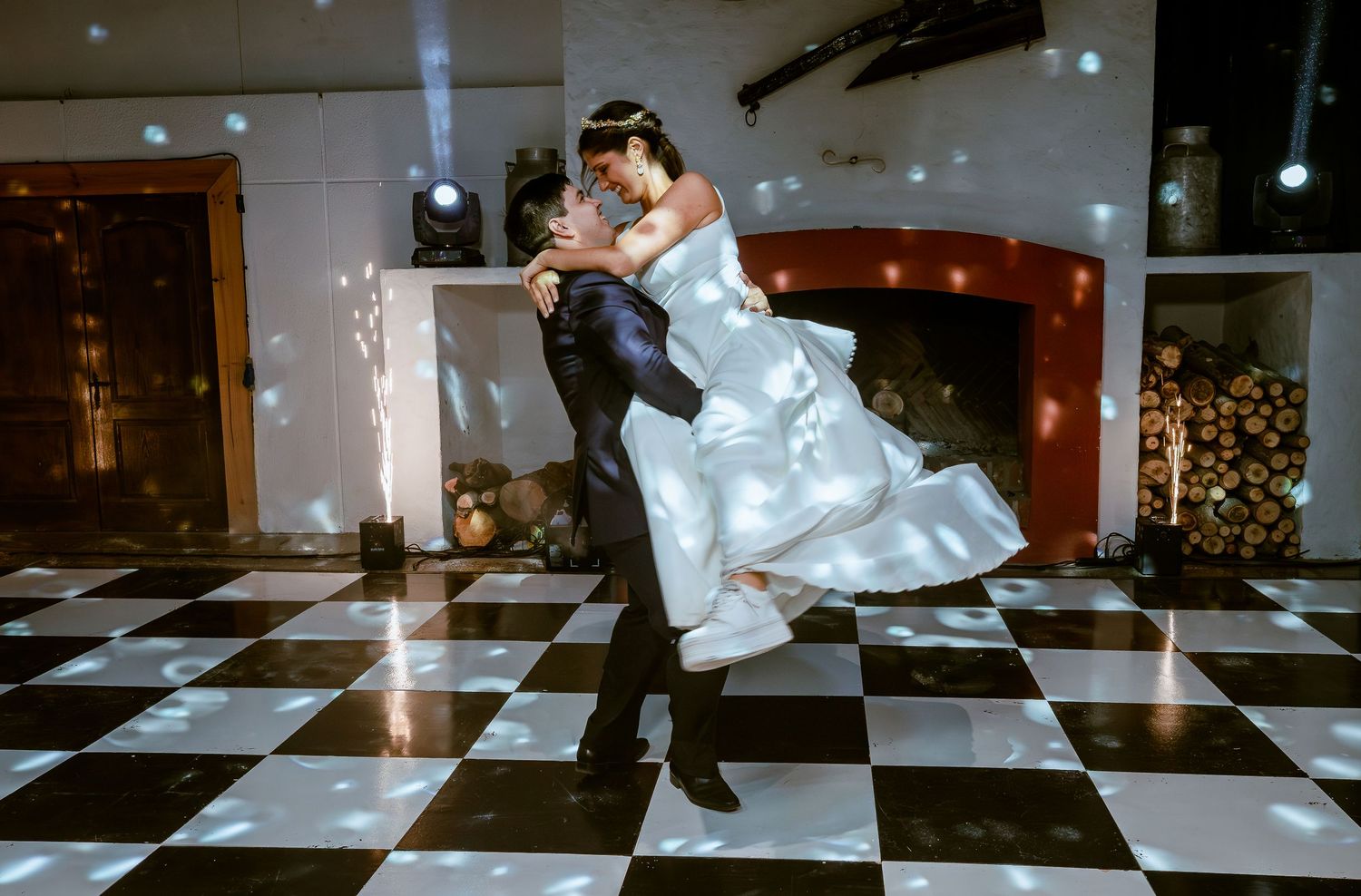 Groom lifts bride during romantic first dance on checkered floor with sparkles.