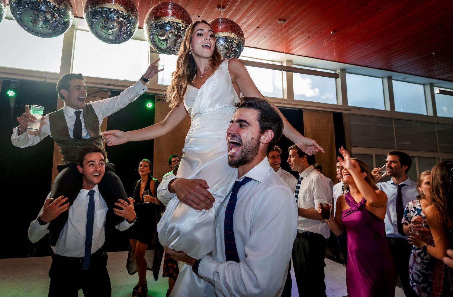 Bride lifted on groom's shoulders as wedding guests cheer and clap on the dance floor.