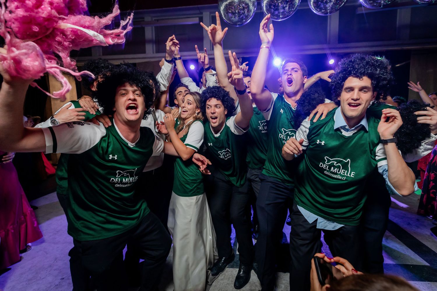 Group of wedding guests wearing matching green sports jerseys celebrate enthusiastically on the dance floor.