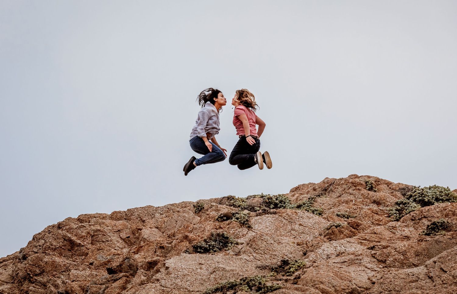 Two women jumping joyfully facing each other above a rocky hillside against clear sky.