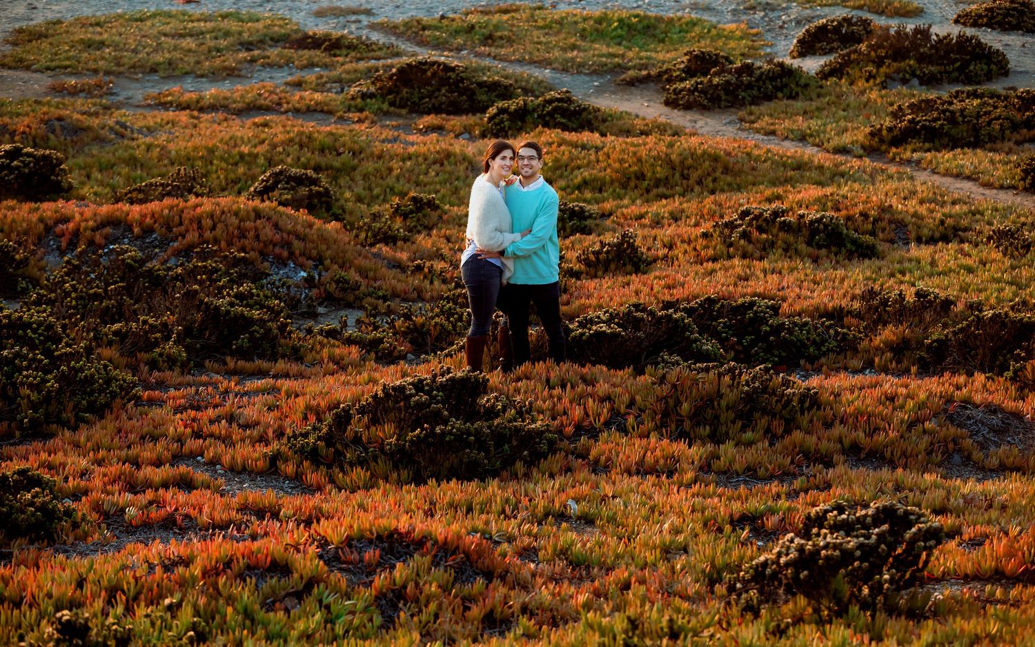 Couple standing together in a colorful field of red and orange coastal plants.