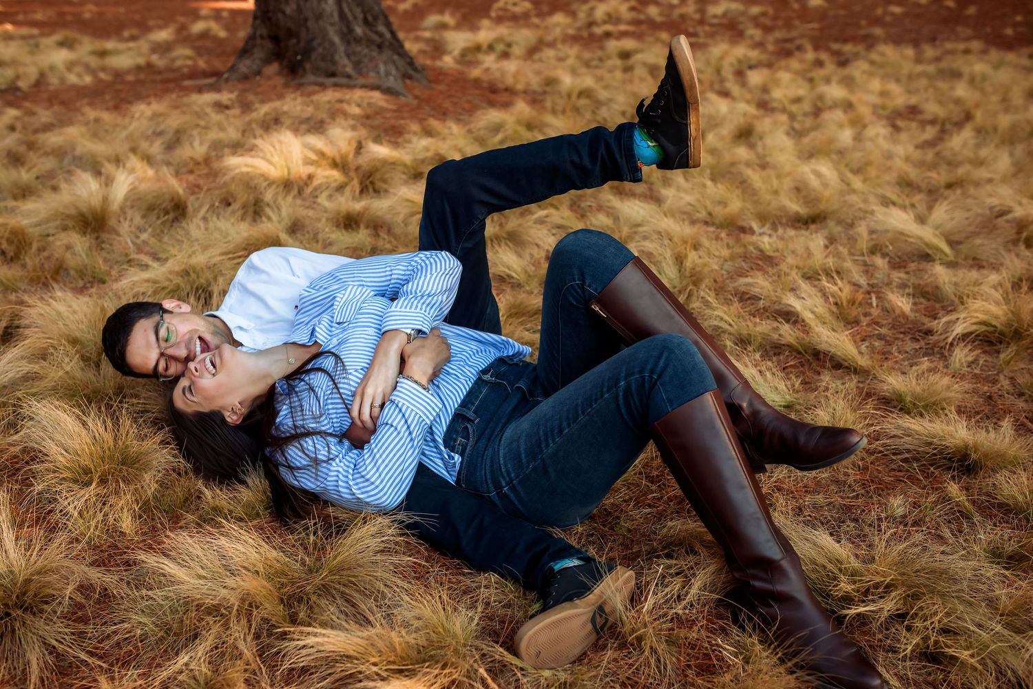 Couple playfully lying together in dry grass field, laughing joyfully.