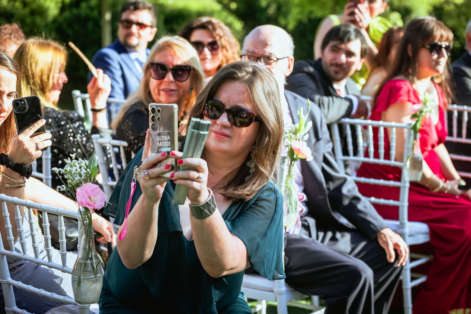 Wedding guest takes selfie on smartphone while seated at outdoor ceremony in sunglasses.