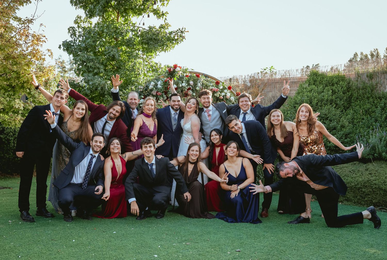 Large wedding party celebrates with bride and groom under floral arch in lush garden setting.