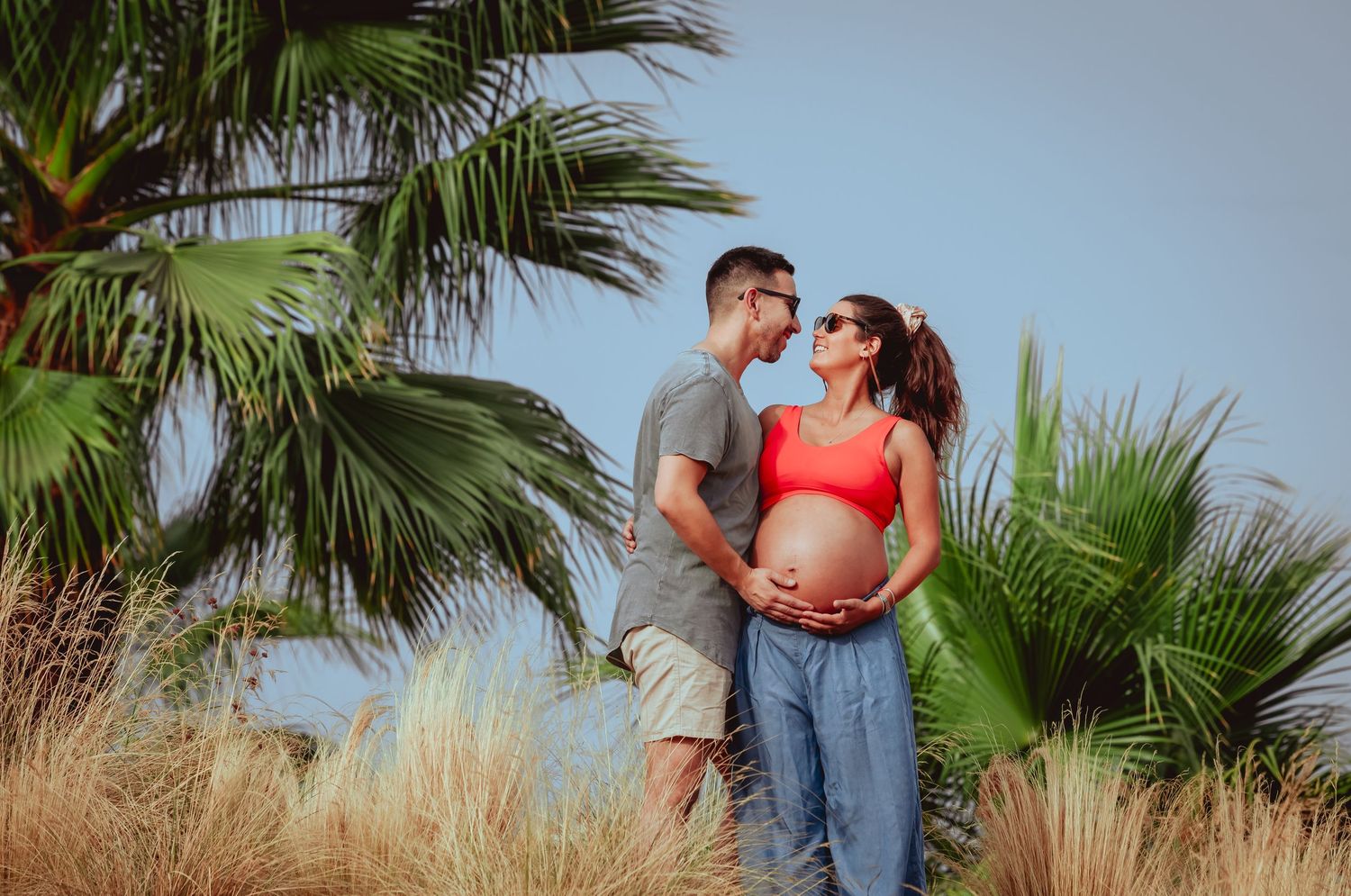 Pregnant couple embracing near tropical palm trees, woman in red crop top and blue pants holding baby bump.
