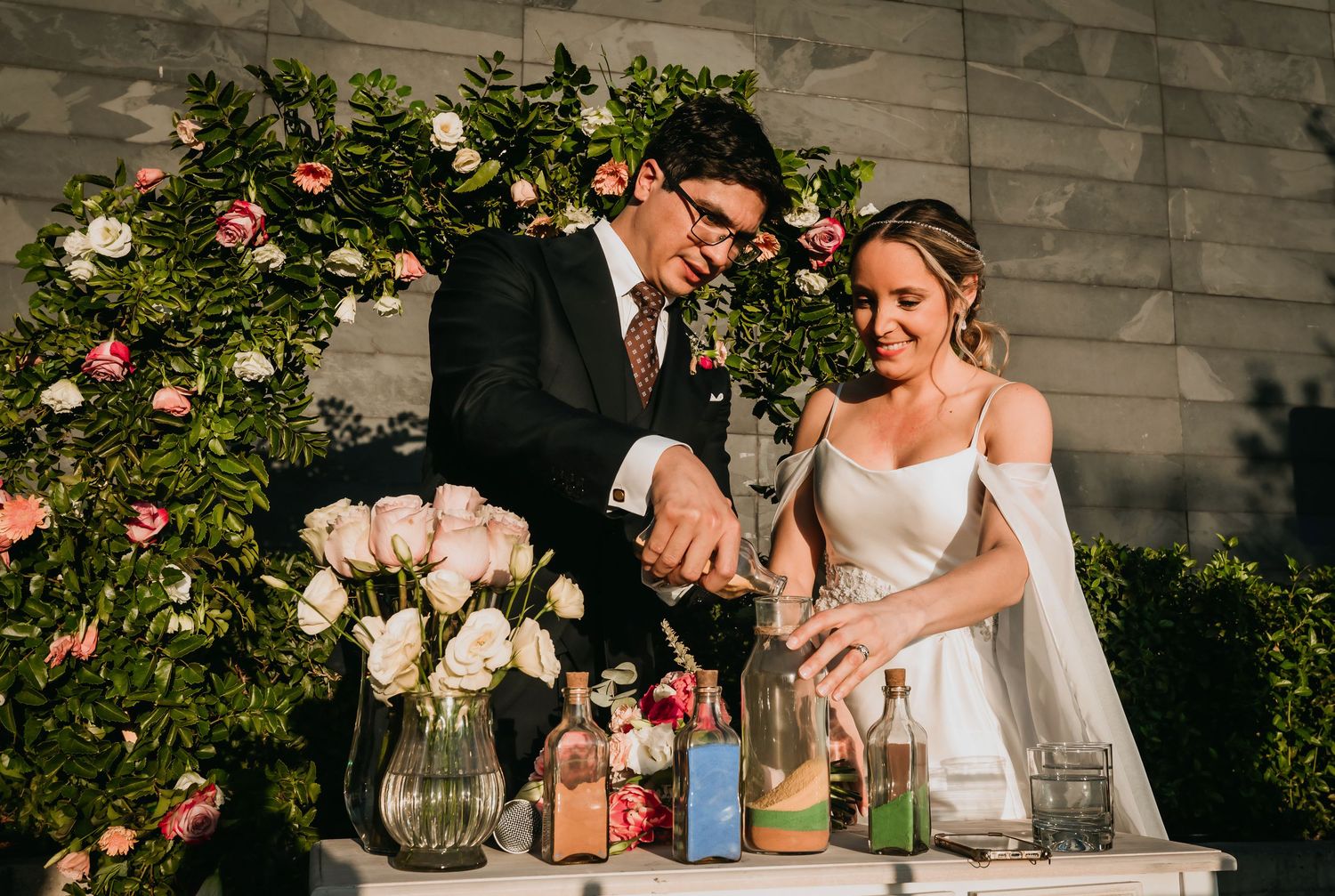 Bride and groom pouring colorful sand in unity ceremony before floral backdrop.