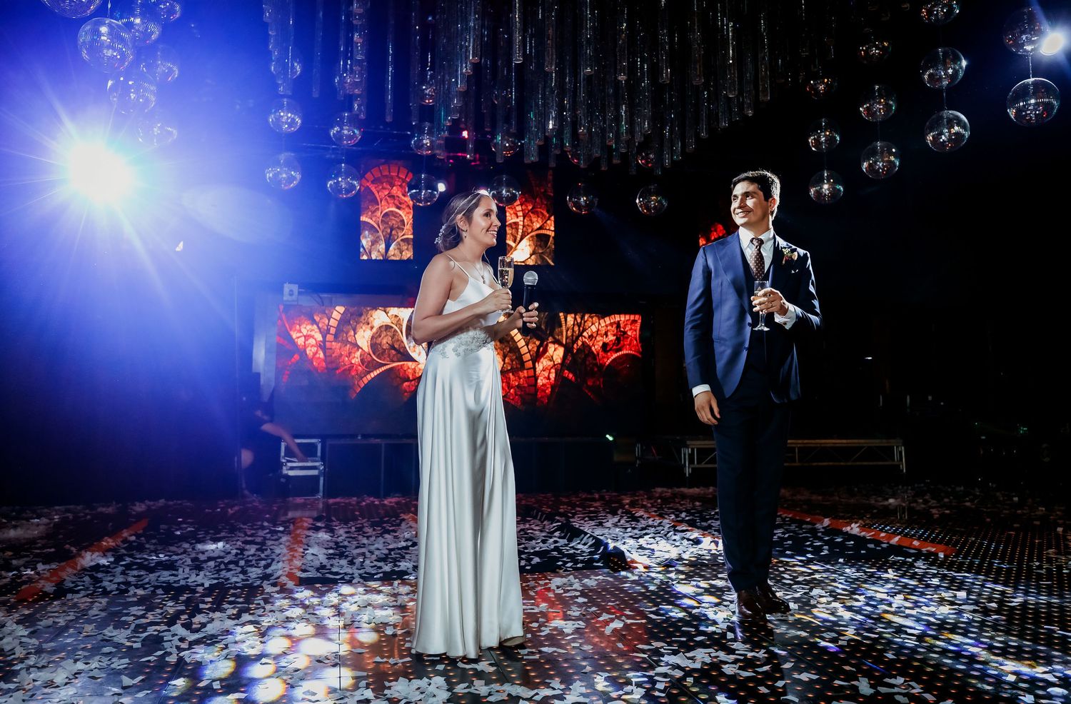 Bride and groom standing elegantly on confetti-covered dance floor with blue stage lighting.