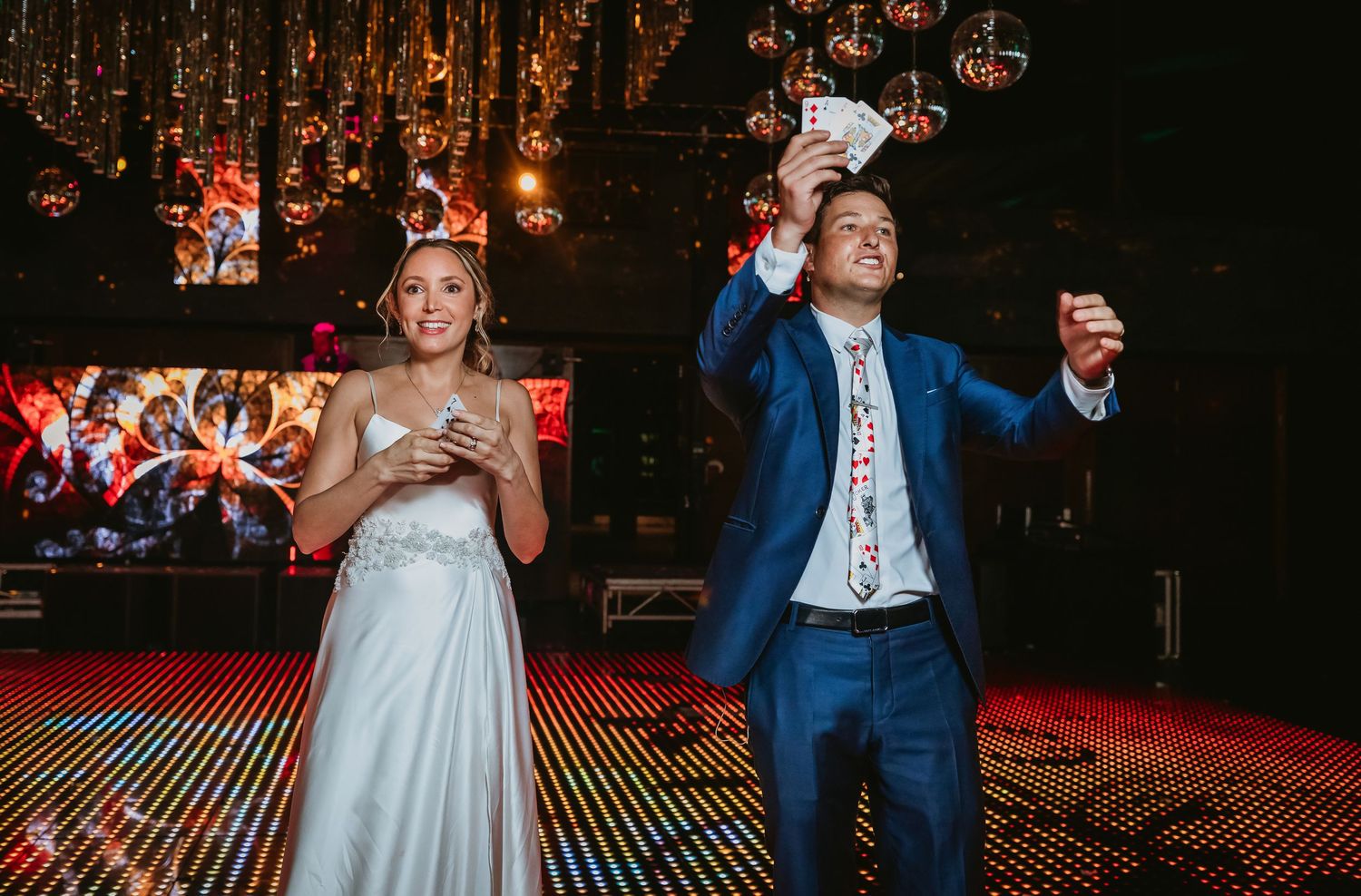 Bride and magician in blue suit performing a card trick on a glittering wedding reception stage.