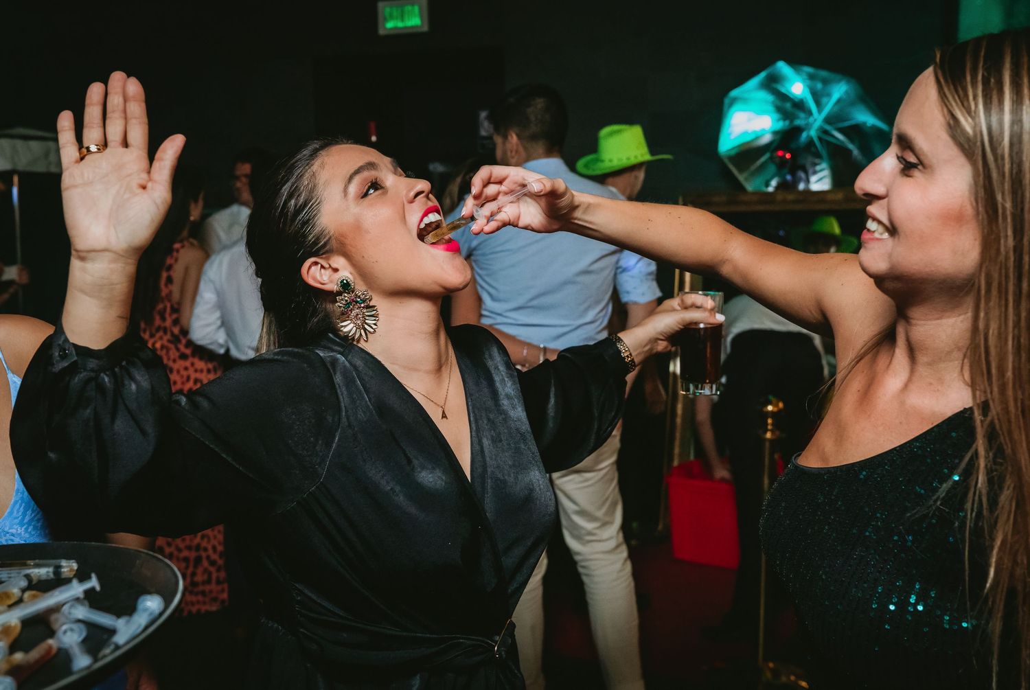 Two women laugh and dance as one feeds the other a syringe shot at a wedding party.