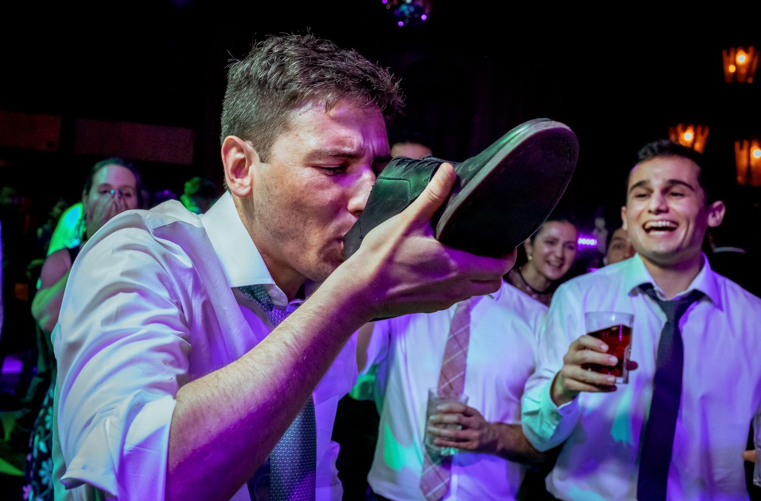 Laughing wedding guest drinks from a shoe while another guest watches and smiles at the reception.