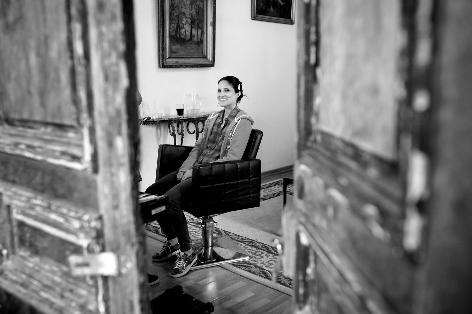 Black and white portrait of woman sitting in vintage chair viewed through ornate wooden door.