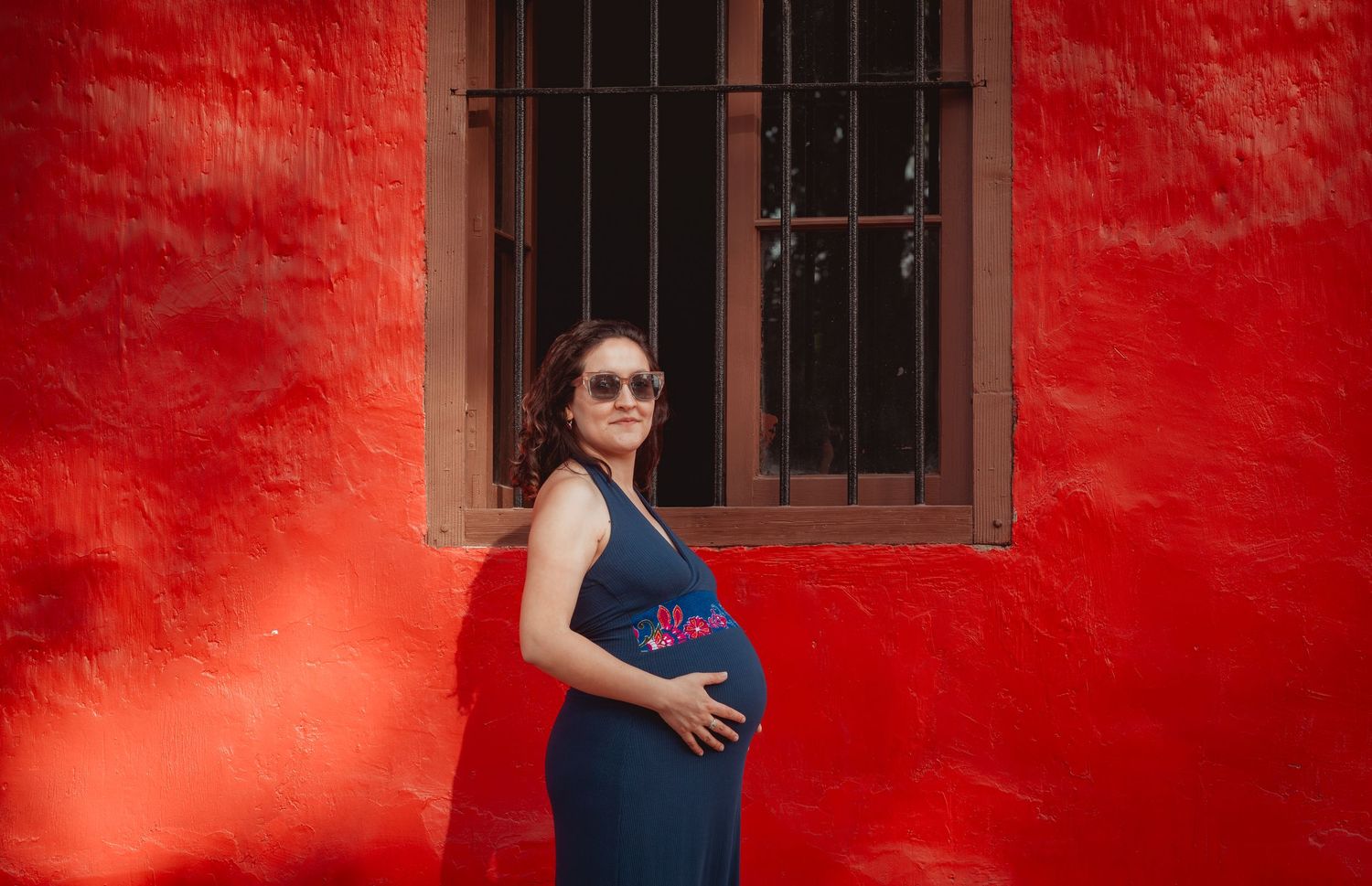 Pregnant woman in sunglasses posing confidently against a bright red wall.