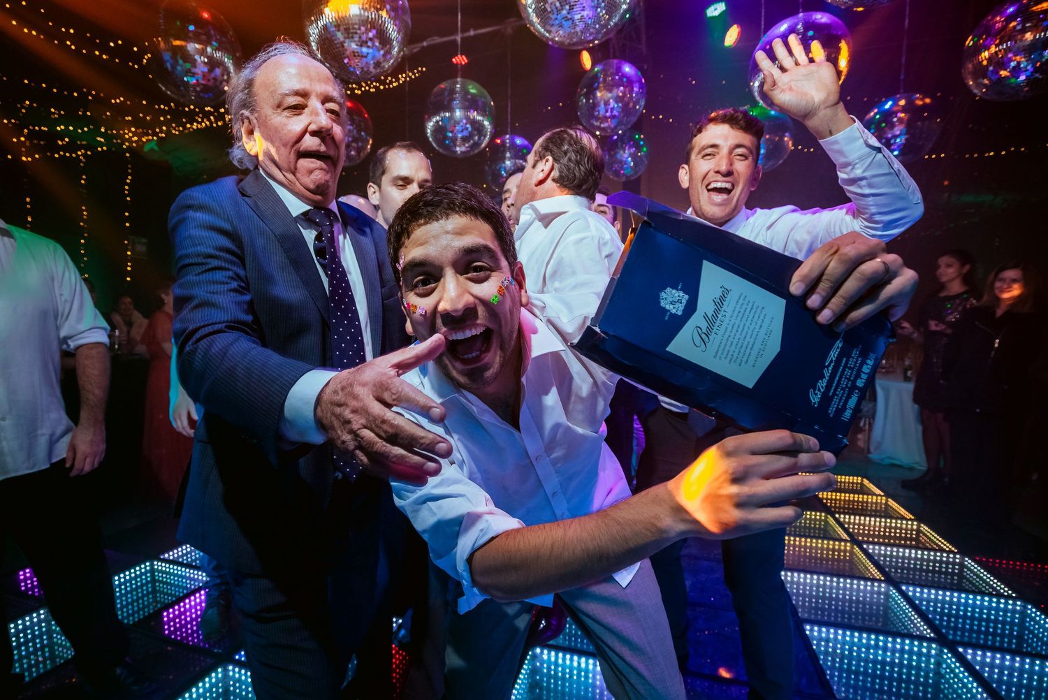 Men celebrating at a disco party, laughing and posing with a gift on a colorful LED dance floor.