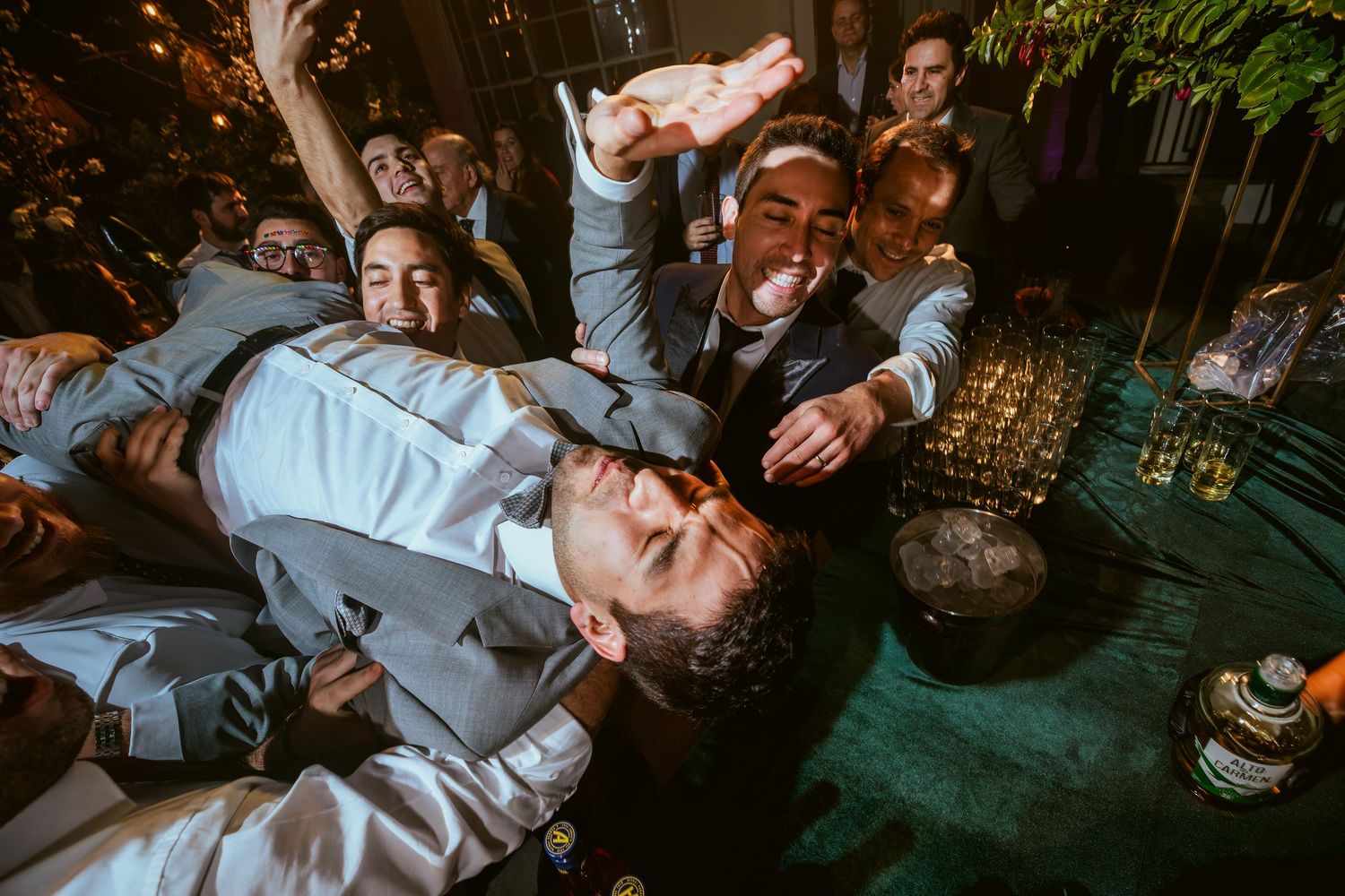 Groomsmen celebrating at a wedding reception, with one being lifted in a lively crowd, holding a drink.