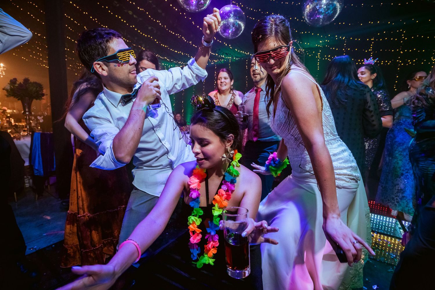 People dancing and celebrating at a colorful party event with disco balls, masks, and Hawaiian leis under vibrant lights.