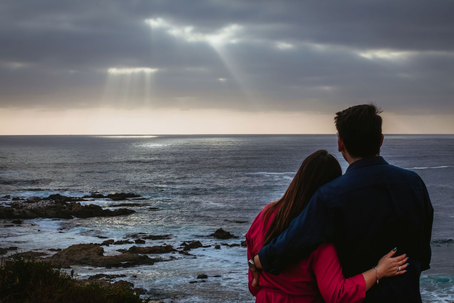 Couple embracing while watching dramatic sunset rays over rocky ocean coastline.