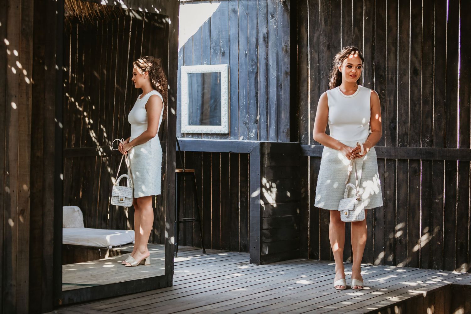 Woman in white dress and skirt reflected in a mirror on a dark wooden deck.