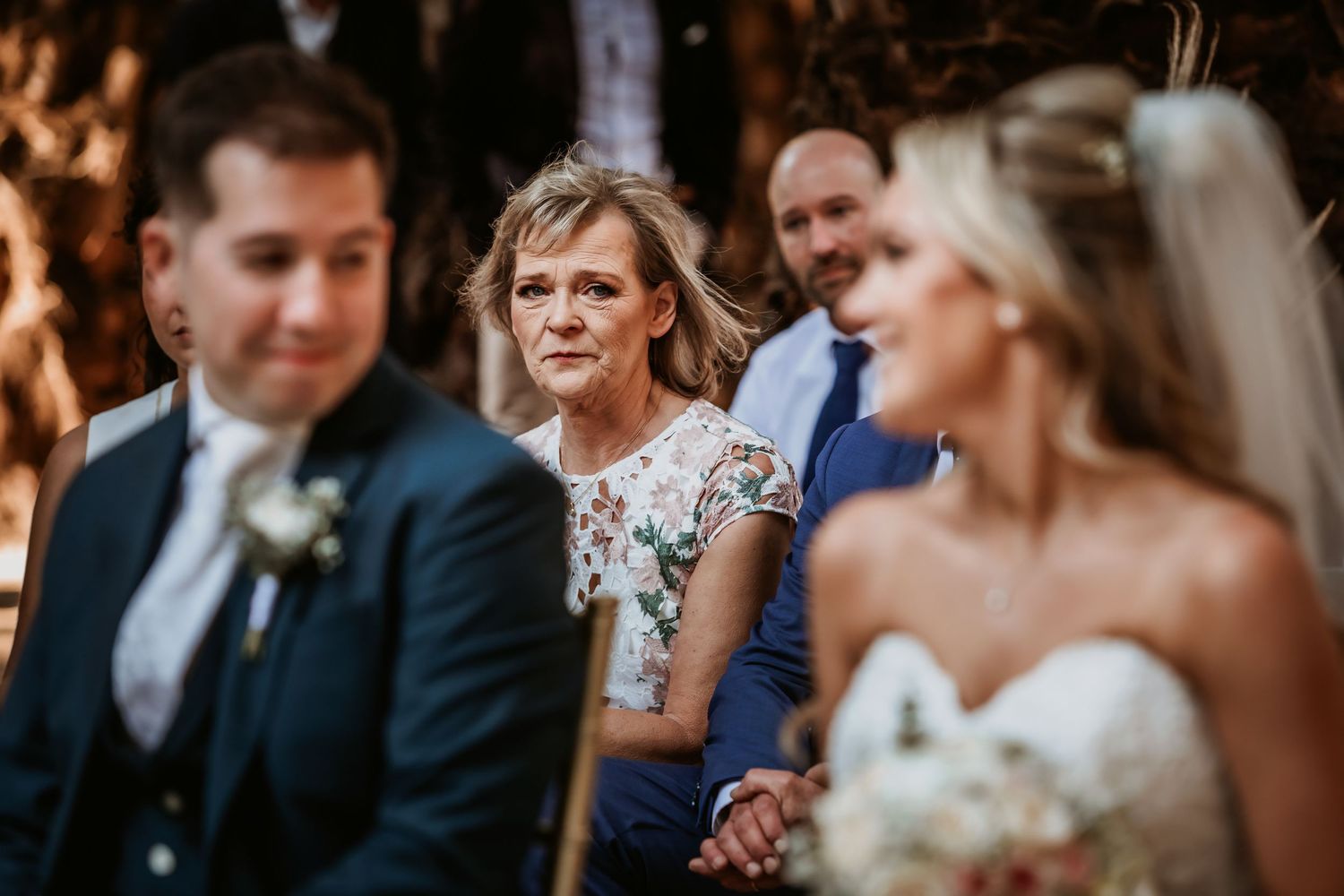 Emotional mother watching wedding ceremony with bride holding bouquet in foreground.