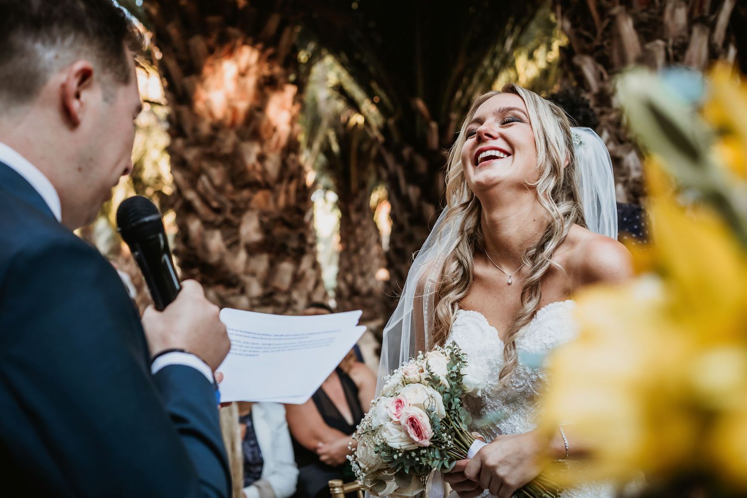 Bride laughs joyfully as her groom reads heartfelt wedding vows at their outdoor palm tree ceremony.