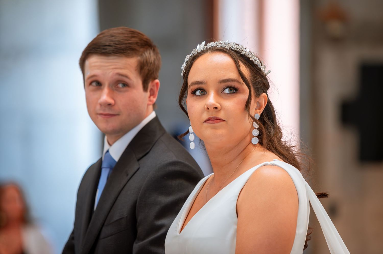 Bride in white gown with tiara and groom in suit stand at church altar.