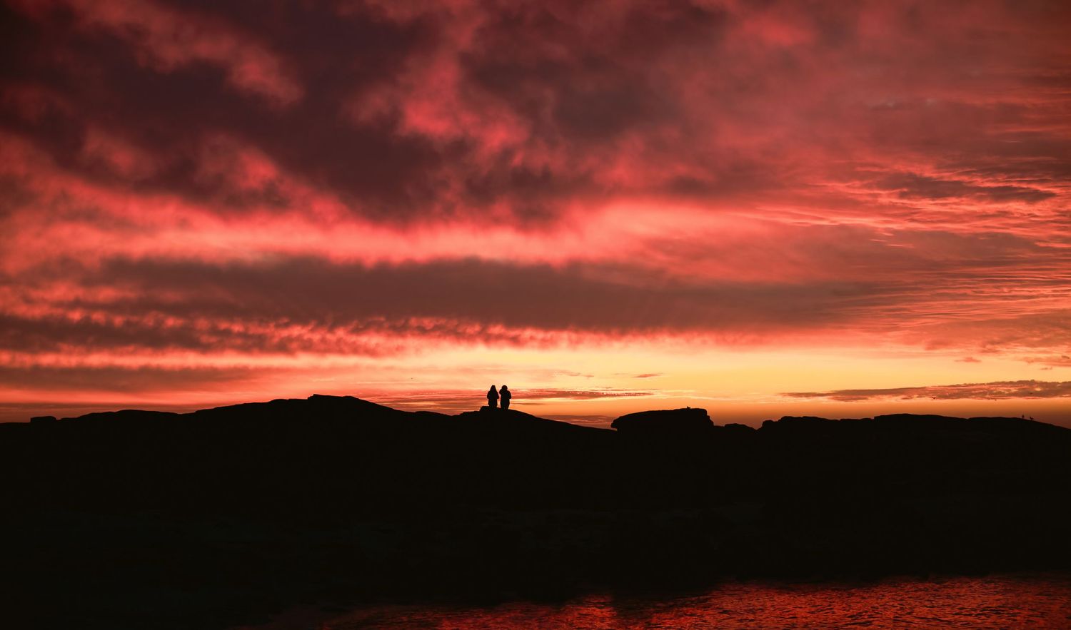 Romantic silhouette of couple standing on rocks against dramatic red sunset sky.