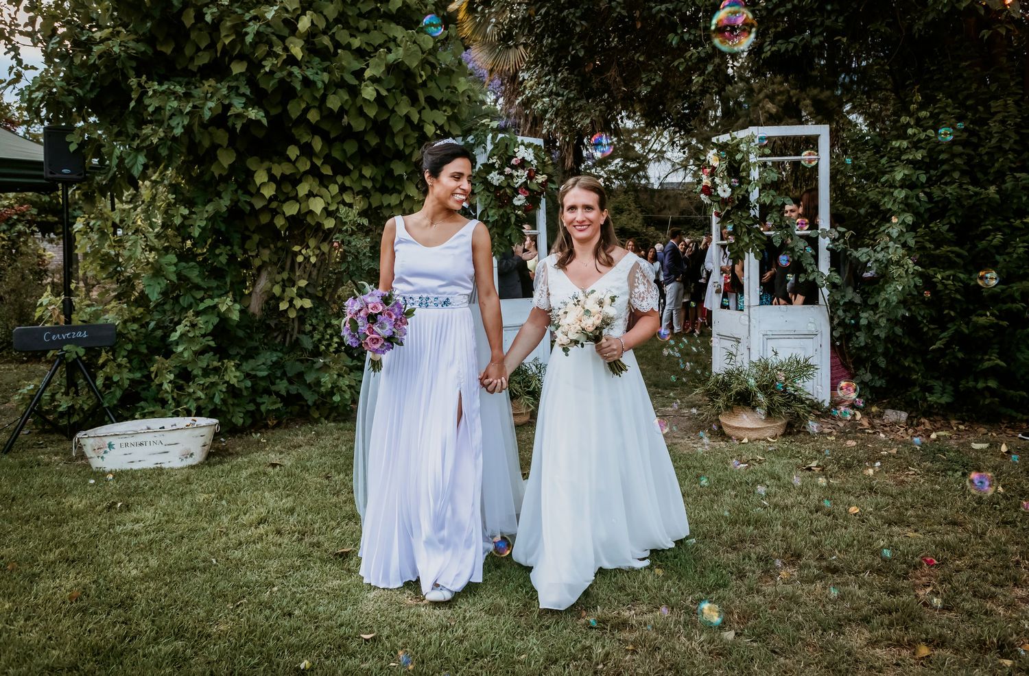 Two brides holding hands and bouquets while posing together at outdoor garden wedding ceremony.