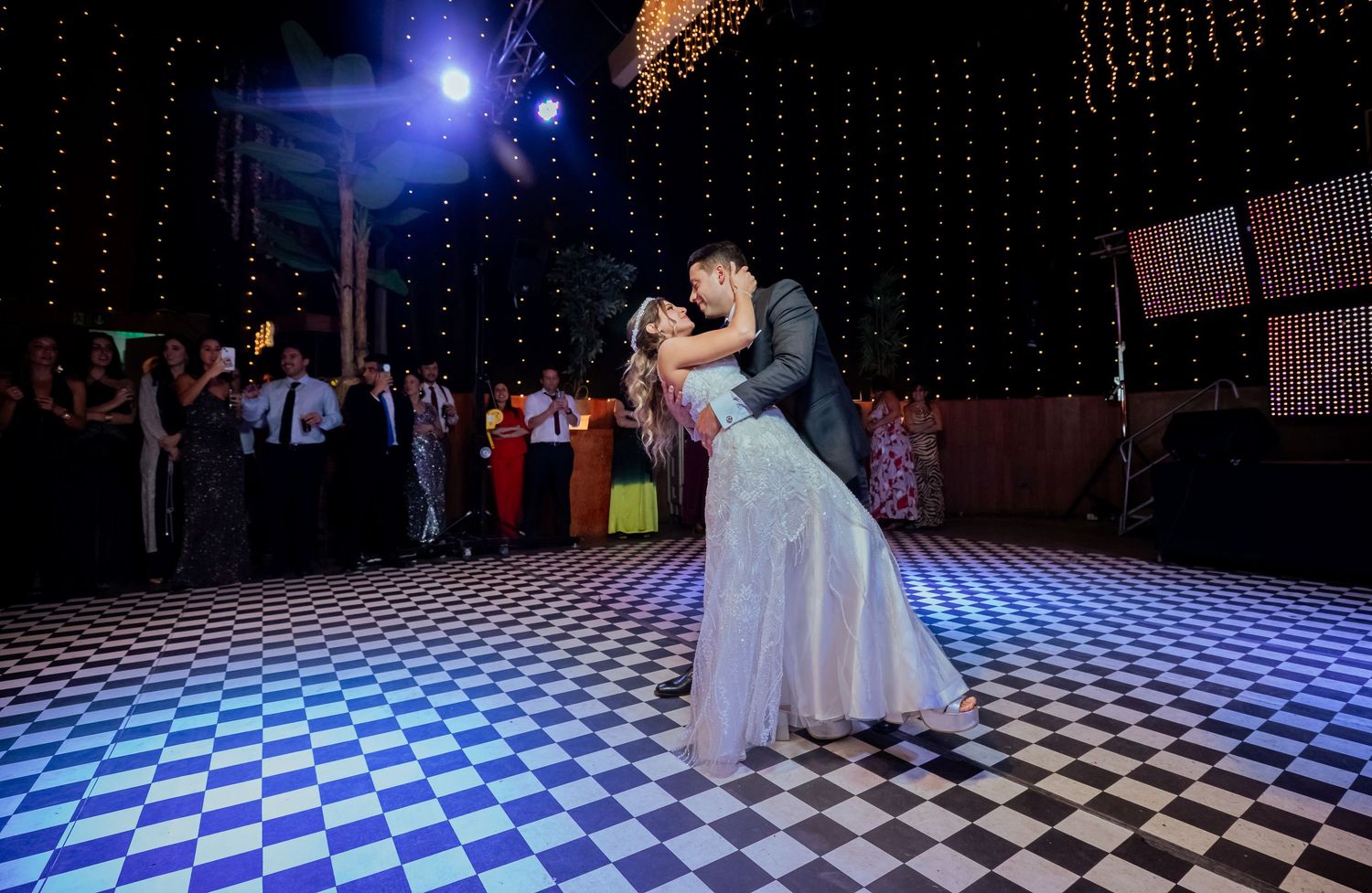 Newlyweds embrace during first dance on checkered floor while guests watch in elegant wedding venue.