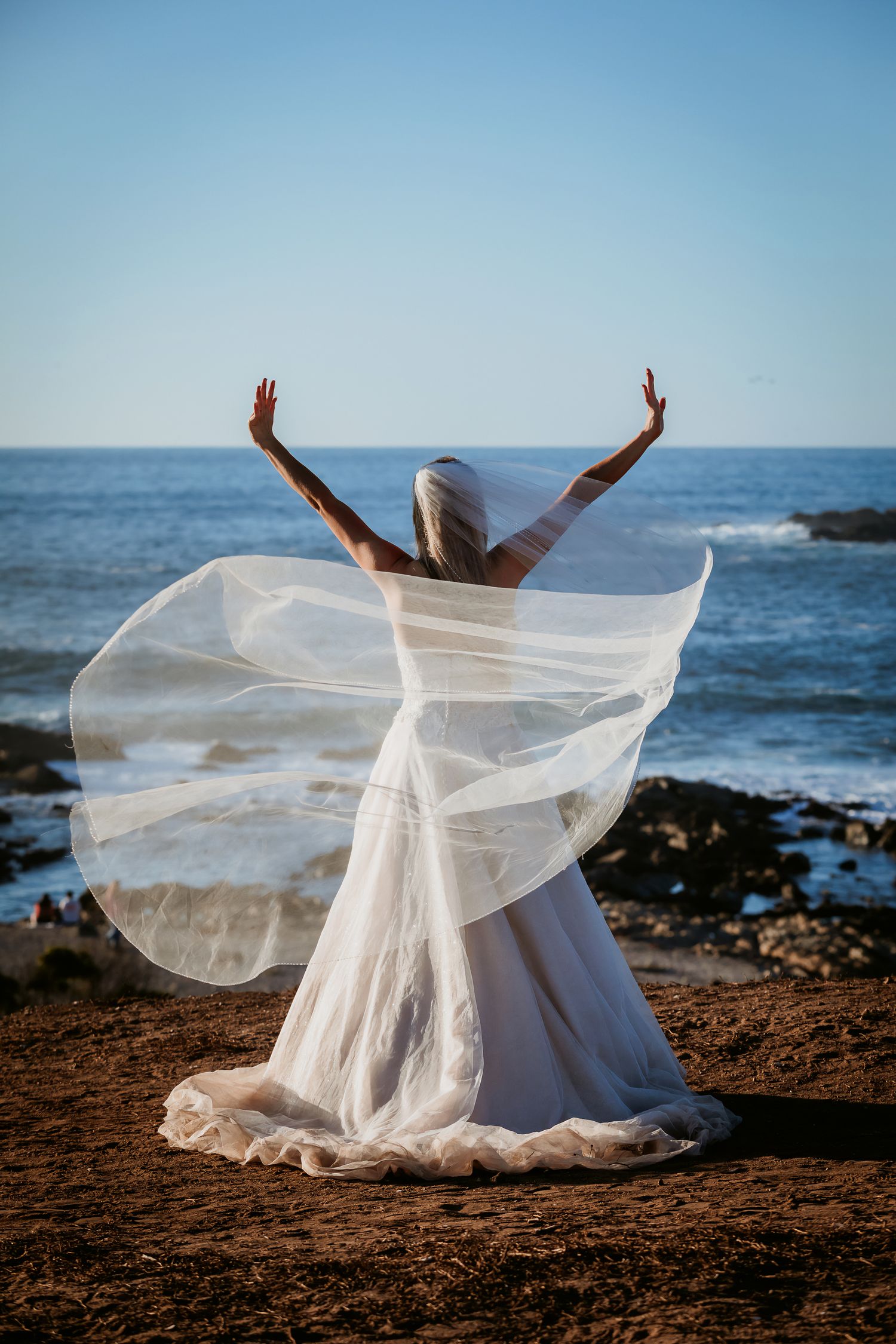 Bride in white gown raises arms toward ocean, veil billowing in wind at rocky coastal shore.