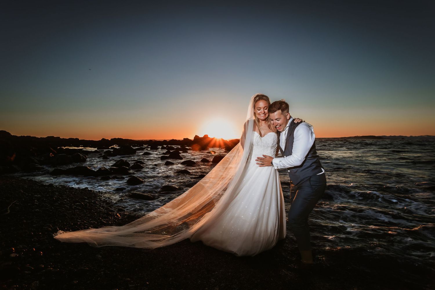Newlyweds sharing a tender moment on rocky beach, bride's veil trailing in sunset light.