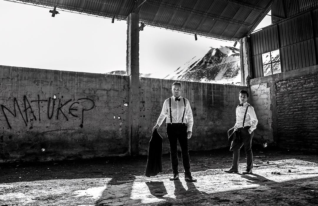 Black and white photo of two grooms in suspenders posing in an abandoned warehouse with graffiti wall.